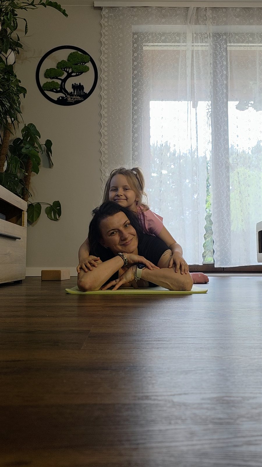 Pani Marta uśmiecha się na macie do jogi, a dziewczynka przytula się do jej pleców